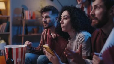 Ciddi erkekler ve dişiler akıllı telefon tutuyorlar ve televizyon ekranına bakıyorlar. Evde oynanan futbol maçı sırasında spor bahislerinin sonuçlarını bekliyorlar ve bundan zevk alıyorlar. Arkadaşlar evde toplanıyor.