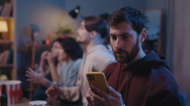 Olumlu sakallı neşeli bir adam futbol maçı sırasında telefonuna mutlu duygularla bakıyor. Evde modern akıllı telefon kullanan yakışıklı traşsız erkek hayranı. Futbol taraftarları konsepti