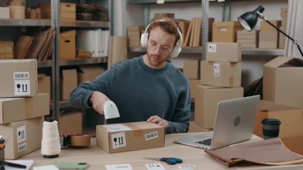 彼のヘッドフォンで音楽を聞きながら 倉庫に座って大きな小包をスキャンジンジャー白人男性 中小企業コンセプト — ストック動画