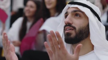 Geleneksel giysiler içinde Arap şeyhi futbol maçında milli takımı desteklerken el kol hareketi yapıyor. Tribünde oturan ve duygusal olarak maç izleyen erkek taraftar..