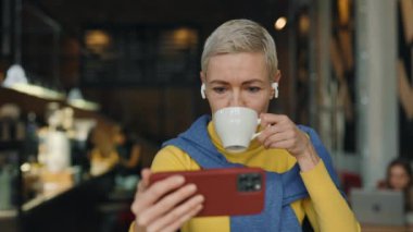 Kafkasyalı olgun bir kadın modern kafede oturuyor, kahve içiyor ve kulaklıkla akıllı telefondan video izliyor. Mavi ve sarı kazaklar giyen çekici bir kadın. İnsanlar ve teknoloji