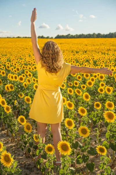 A woman with her back to a field of sunflowers with her arms raised in a yellow dress.