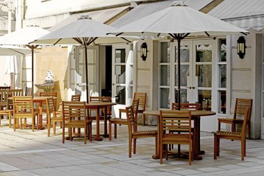 Ahşap kafe masaları ve şemsiyeli sandalyeler, çoğunlukla beyaz olarak tasarlanan avlu hizasında.