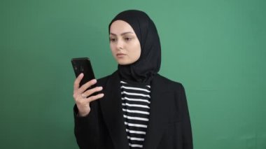 Yeşil telefon ekranına bakan Müslüman bir kadın, izole edilmiş yeşil arka plan önünde akıllı telefon gösteren genç bir kız, gülümseme.