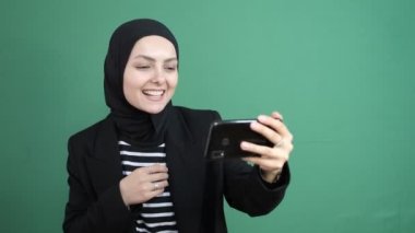 Müslüman bir kadının yeşil ekranın önünde görüntülü araması, kız gülüyor, genç bir kızın neşeli bir şekilde arkadaş telefonuyla konuşması