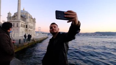 Boğazın tarihi binası Ortakoy Camii ile aynı meydanda yer almaktadır. Genç adam, Boğazın ve arkasındaki binanın manzarasıyla fotoğraf çekmektedir.