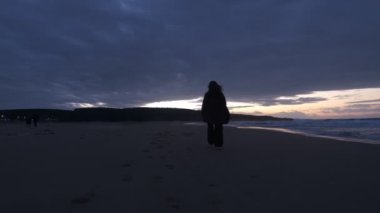 Güneşin batışına doğru adım atarak ve rüzgarlı havada deniz kenarında el sallayarak yalnız yürüyen bir kadın.