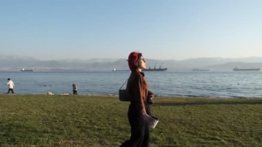 Genç ve güzel bir kadın güneşli bir pazar günü körfezdeki parkta vakit geçiriyor. Sahil kenti parkında, arkada gemiler var.