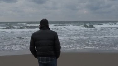 Denizin kenarında durup çevreyi izleyen genç adam, manzaraya bakmak için kameranın önünden geçen genç adam, denizde taş sektirerek zaman harcıyor.