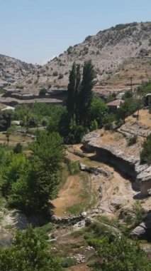 Yeşil bir vadide akan bir dere ve yamaçta inşa edilmiş bir taş bina selvi ağaçları ve uçak ağaçlarıyla kaplı bir dere.