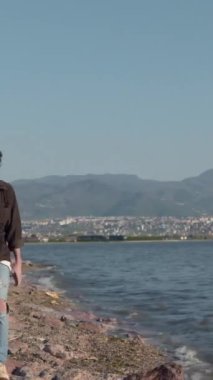 Kulağında kulaklık olan genç bir adam deniz kenarında yürüyor. Elinde telefonla sahilde tek başına yürüyor.