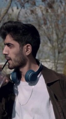 Genç adam parkta dikilip içkisini içiyor, kulaklıkları boynunda bir ağaca yaslanmış soğuk kahve içiyor.