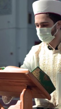 Yüzünde sağlık maskesi olan cami öğretmeni Kuran okuyor. Masada duruyor, imam kutsal kitaba bakıyor.