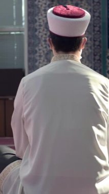 Cami öğretmeninin tek başına oturduğu ve arkası dönük camide, cüppeli genç bir adamın mescitte oturduğu resmi.