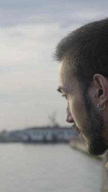 Küpeli genç bir adam kumsalda oturmuş denizi düşünüyor güneşli bir günde denizi seyrediyor.