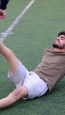 Ayakta duran genç adam halıya uzanmış bacaklarını hareket ettiriyor, genç insanlar açık havada halıda spor yapıyor.