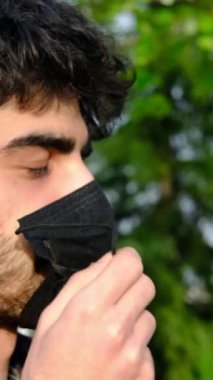 Maskeli genç adam yeşil ağaçlarla arka planda duruyor maskesini çıkarıyor ve içme suyu içiyor, sakallı genç bir adam su içiyor.