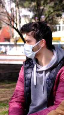 Yüzü maskeli genç bir adamın, yanında oturan iki arkadaşıyla konuşurken, genç insanların şehir parkında oturup konuştuğu farklı bir açı çekimi.