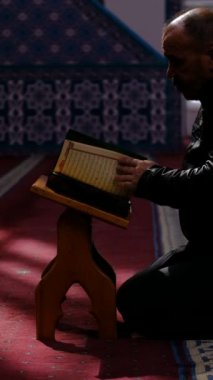 Camide dizlerinin üzerinde oturan yaşlı adam Kuran okuyor, sayfaları çeviriyor ve Müslümanlar İslam 'ın kutsal kitabını okuyorlar.