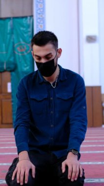 Genç adam dua ederken sağa sola başını sallıyor, caminin tente kapısının önünde oturan adam dua ediyor.