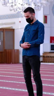 Yüzünde tıbbi maske olan müslüman bir gencin camide dua edişinin farklı açılardan çekimi. İslam 'a inananlar Tanrı' ya dua ediyorlar.