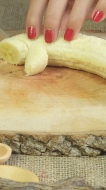 Image of woman hand slicing yellow banana on wooden cutting board, fruit preparation on thematic natural products table, slicing process on wooden presentation plate