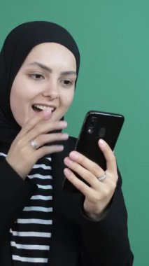 Müslüman kadın telefon şokuna bakıyor, yeşil ekranın önünde gülen siyah tesettürlü kız, genç insan telefon ekranını gördü çok mutlu.