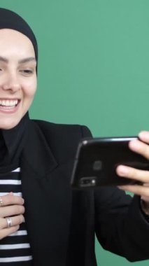 Müslüman bir kadının yeşil ekranın önünde görüntülü araması, kız gülüyor, genç bir kızın neşeli bir şekilde arkadaş telefonuyla konuşması