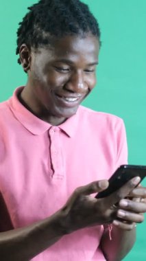 Siyahi bir adam heyecanlı bir şekilde mesaj atıyor, gülümseyen siyah bir erkek telefonda mesajlaşıyor, telefonunuzla eğleniyor, yeşil ekranın önünde vücut dilini ve yüz ifadelerini gösteriyor.
