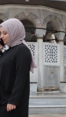 Cami avlusunda tesettürlü bir kadın telefonda konuşuyor, cami binalarının önünde telefon görüşmesi yapıyor ve fıskiyenin arkasında da fıskiye var.