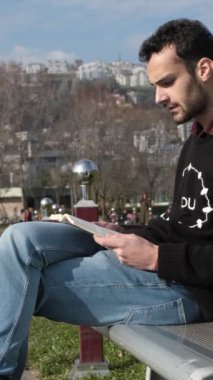 Güneşli bir günde beyaz bankta kitap okuyan yalnız bir adamın görüntüsü, rahatlama aktivitesi bankta oturup güneşin tadını çıkarması, parkta yeşil çimlerle yapılan aktivite.
