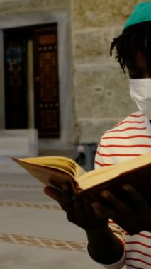 Yüzünde sağlık maskesi olan Afrikalı bir genç camide oturmuş Kuran okuyor, Müslüman erkekler camiye gidiyor.