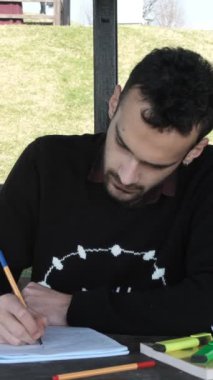 Park 'ta ders çalışan yalnız bir adamın görüntüsü, Arbour' da ödev yapan genç bir adam, ahşap bankta çalışma kağıdını tamamlamış, güneşli bir günde parkta sınavlara hazırlanıyor.