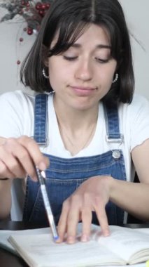 Kalemle çalışırken sıkılan, çalışma ortamında kitap ve kalemlerle oynayan, sıkı çalışmaktan bıkmış genç bir kız.