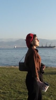 Genç ve güzel bir kadın güneşli bir pazar günü körfezdeki parkta vakit geçiriyor. Sahil kenti parkında, arkada gemiler var.
