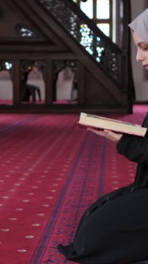 Genç Müslüman kadın caminin içinde Kuran okuyor, ahşap kürsüde el-Kuran okuyor ve içeride diz çöküp dua ediyor.