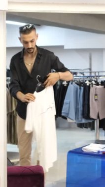 Shopping boy trying on t-shirt, looking at the shirt he likes in the mirror, try on white t-shirt, man trying product in cabin to buy