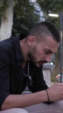 Alışveriş uygulaması kullanan bir adam, telefonla alışveriş yapan bir genç, dijital alışveriş yapan, bankın dışında alışveriş yapan, bankta kırmızı yeşil bir alışveriş çantası olan bir adam.