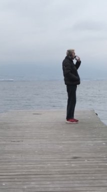 Sonbaharda ahşap rıhtımda kahve içen genç adam, bulutlu bir havada limanda açık hava aktivitesi olarak vakit geçiriyor.