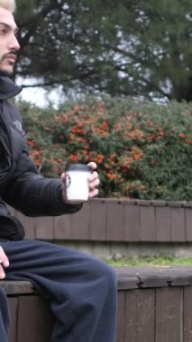 Genç bir adam parkta ağaçlarla oturuyor, eğleniyor, bankta sigara içiyor ve kahve içiyor, sonbaharda tek başına açık hava aktivitesi yapıyor.