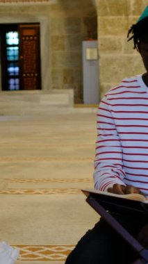 Afrikalı siyahi Müslüman ergen camide oturmuş, masada dini bir kitap okuyor, Müslüman erkekler camiye gidiyor.