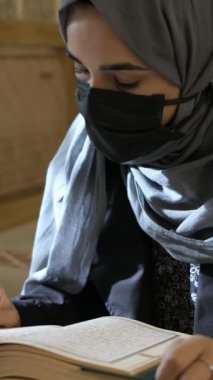 Maskeli Müslüman kadın Kuran okuyor, genç kız ahşap kürsüde Kuran okuyor, İslam kurallarına göre ibadet ediyor.