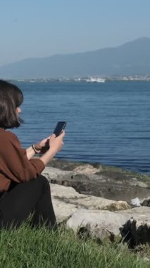 Deniz manzarası karşısında oturan güzel kız kulaklıkla müzik dinliyor, kulaklık takıyor ve çevreyi seyrediyor.