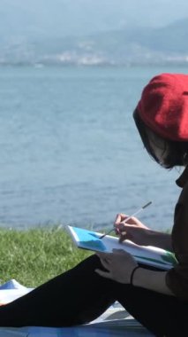 Deniz manzarası ve resim karşısında oturan genç kadın ressam, deniz kenarındaki çimenlerde oturan ve tuvale resim çizen kadın.