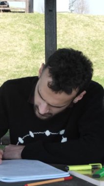 Park 'ta ders çalışan yalnız bir adamın görüntüsü, Arbour' da ödev yapan genç bir adam, ahşap bankta çalışma kağıdını tamamlamış, güneşli bir günde parkta sınavlara hazırlanıyor.