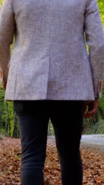 Ceketli genç bir adam ormanda kahverengi yapraklar üzerinde yürüyor, doğada yürüyen bir adam, ormanda sararmış yapraklar yere dökülmüş.