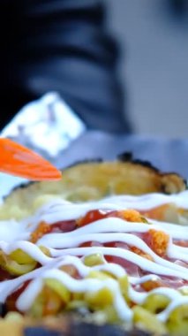 Genç bir adam sokakta oturmuş fırında patates yiyor, çeşitli soslarla süslenmiş bir sokak yemeği aperatifini tadıyor.