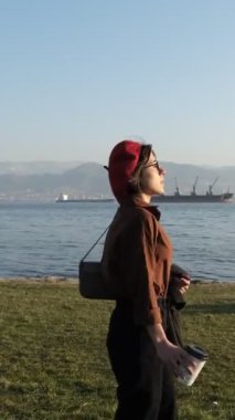 Genç ve güzel bir kadın güneşli bir pazar günü körfezdeki parkta vakit geçiriyor. Sahil kenti parkında, arkada gemiler var.