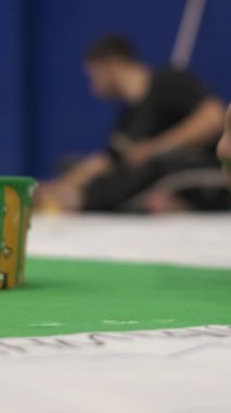 Bir el yeşil boyayı boya açısıyla dağıtıyor, yerdeki bezi farklı yöntemlerle fırça hareketiyle boyuyor.