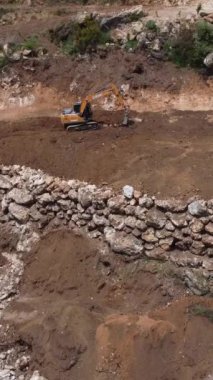 Sürüngen inşaat makinesinin havadan görünüşü eğimli arazide duvarlar inşa ederken bir eziciyle kayaları eziyor.